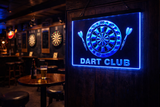 Neon sign for a dart club in a bar setting with dartboards on the wall.