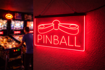 Red Pinball LED sign with flippers and ball hanging on a wall beside a row of pinball machines